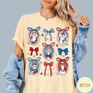 May include: A light yellow t-shirt featuring a patriotic design. The design includes cartoon geese in red, white, and blue outfits, surrounded by bows and floral accents. The shirt has a relaxed fit.