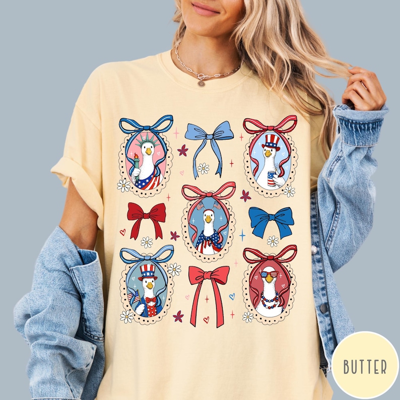 May include: A light yellow t-shirt featuring a patriotic design. The design includes cartoon geese in red, white, and blue outfits, surrounded by bows and floral accents. The shirt has a relaxed fit.