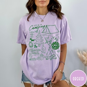 Może przedstawiać: Jasnofioletowa koszulka z zieloną grafiką o tematyce kempingowej. Projekt zawiera tekst "Let's Go Camping" i "Adventure Awaits", a także ilustracje namiotu, ogniska i innych element&oacute;w kempingowych.