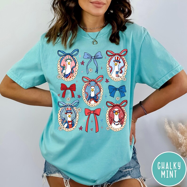 May include: A light blue t-shirt featuring a grid of cartoon geese in patriotic outfits, each framed by a decorative border and bow. The geese wear hats, sunglasses, and bow ties in red, white, and blue. The shirt has a relaxed fit.