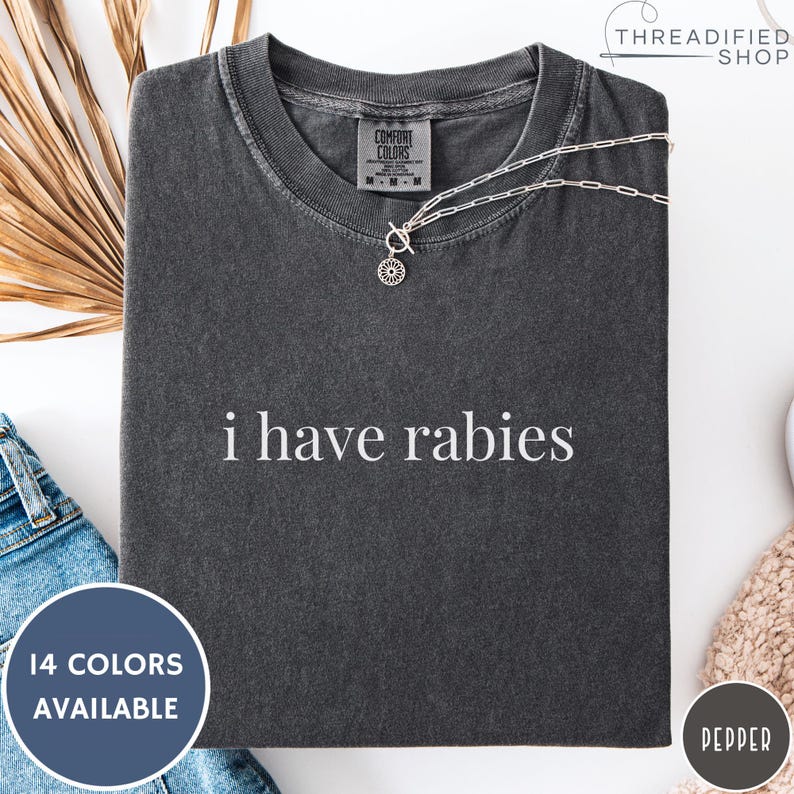 May include: Dark gray Comfort Colors t-shirt with the phrase "i have rabies" in white. A silver necklace with a circular pendant is on the shirt. The shirt is displayed with a blue circle that says "14 COLORS AVAILABLE".