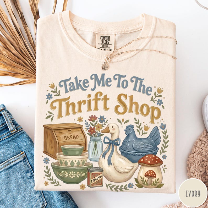 Op de afbeelding: Ivoor kleurig T-shirt met de tekst "Take Me To The Thrift Shop" in blauw en goud. De afbeelding bevat een brooddoos, kommen, een eend, een kip en een paddenstoel.