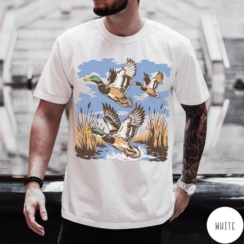 May include: White t-shirt with a graphic of three ducks flying over water and reeds. The ducks are depicted with brown, white, and green details, set against a blue sky. The shirt has a classic crew neck.