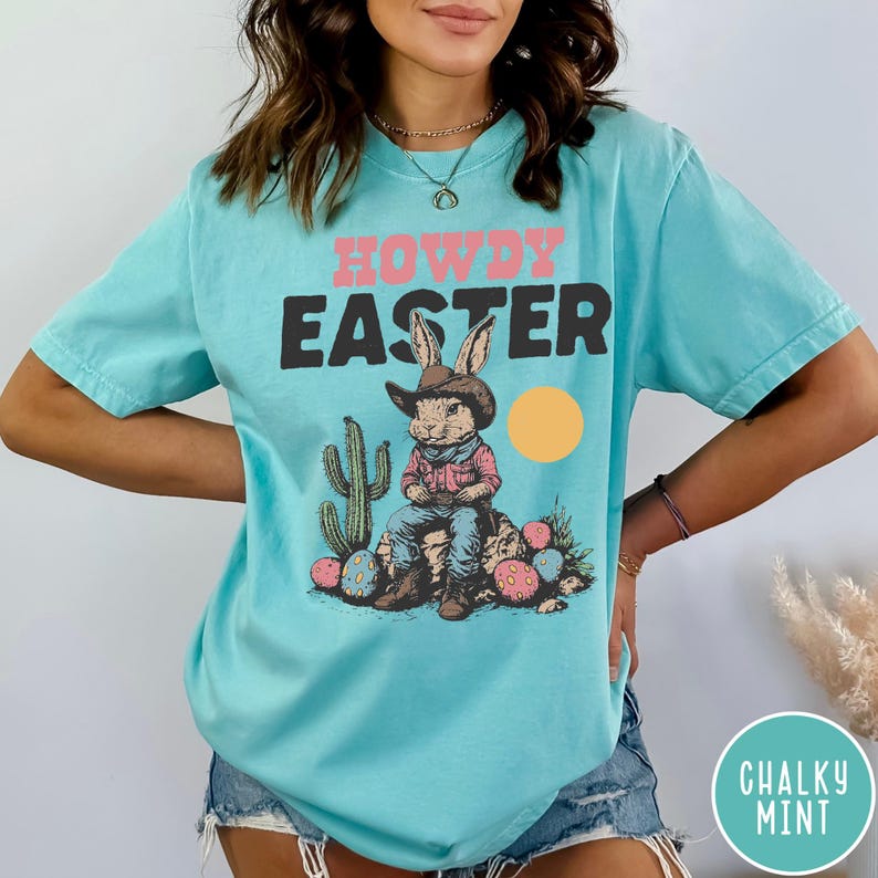 Puede incluir: Camiseta azul claro con las palabras "HOWDY EASTER" en rosa y negro. El gr&aacute;fico presenta un conejo con sombrero de vaquero, sentado en una roca con huevos de Pascua y un cactus. Tambi&eacute;n hay un sol amarillo.