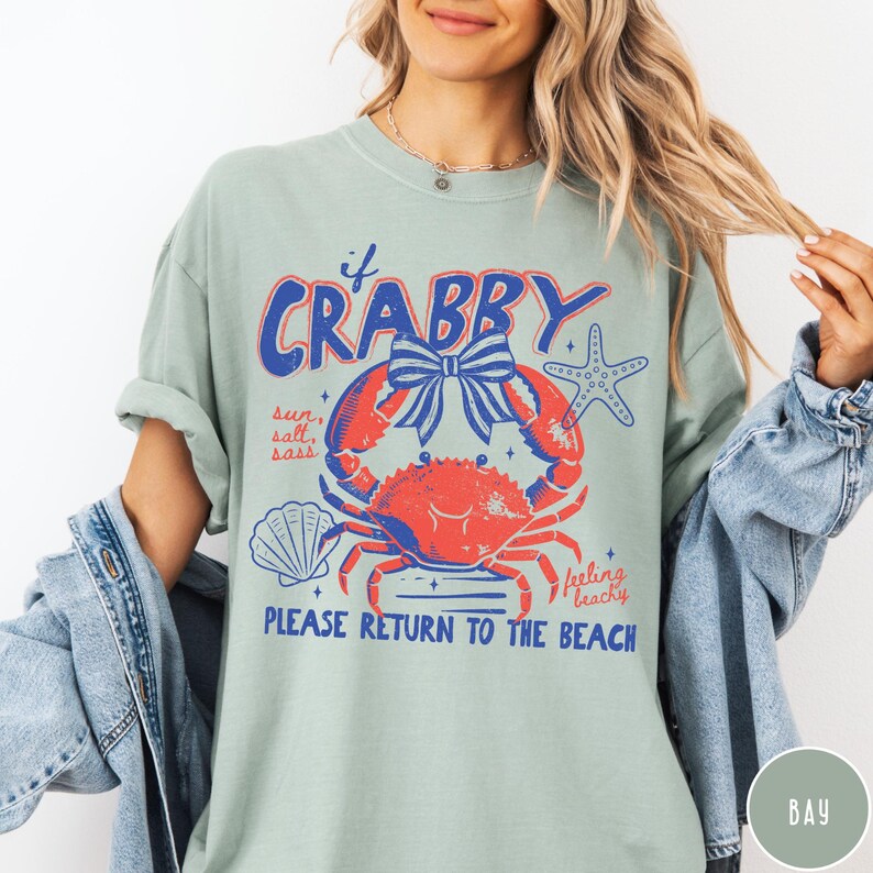 Pode incluir: Camiseta verde s&aacute;lvia com um gr&aacute;fico com um caranguejo vermelho, um la&ccedil;o azul e uma estrela do mar. O texto inclui "If Crabby" e "Please Return to the Beach". Um design divertido com tema de praia.