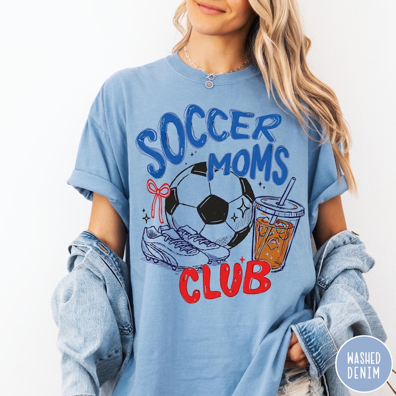 May include: Light blue t-shirt with the words "Soccer Moms Club" in blue and red. The graphic includes a soccer ball, soccer cleats, and a beverage. The shirt is made of a soft material and has short sleeves.
