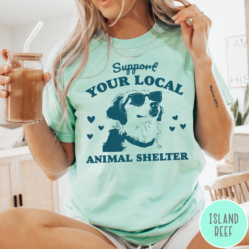 May include: A light blue t-shirt with the text "Support Your Local Animal Shelter" in dark blue. A cartoon dog wearing sunglasses is in the center of the text.