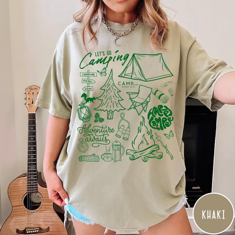 Może przedstawiać: Khaki koszulka z zieloną grafiką o tematyce kempingowej. Projekt zawiera tekst "Let's Go Camping" i "Adventure Awaits", a także ilustracje namiotu, ogniska i innych element&oacute;w kempingowych.