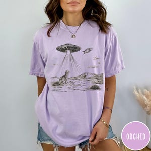 Pode incluir: Uma camiseta lavanda com um gr&aacute;fico de um gato olhando para cima para um OVNI. O OVNI est&aacute; emitindo raios de luz. O texto "ORCHID" &eacute; vis&iacute;vel no canto inferior direito da imagem.