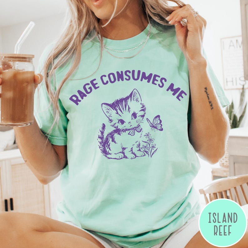 Puede incluir: Una camiseta verde claro con el texto "Rage Consumes Me" en letras moradas. Un gatito de dibujos animados con una mariposa est&aacute; impreso en la camiseta.
