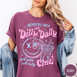 May include: Berry-colored t-shirt with rolled-up sleeves. The front features a graphic with the text "MEMBERS ONLY the Dilly Dally Club" and a smiley face design with a drink. The phrase "half dilly half dally" is also included.