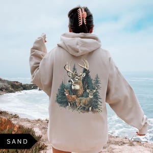 Op de afbeelding: Zandkleurige hoodie met een gedetailleerde afbeelding van herten in een bos. Het ontwerp toont een hert met groot gewei en twee kleinere herten. De hoodie heeft een capuchon met trekkoord en lange mouwen. Het woord "SAND" staat linksonder.