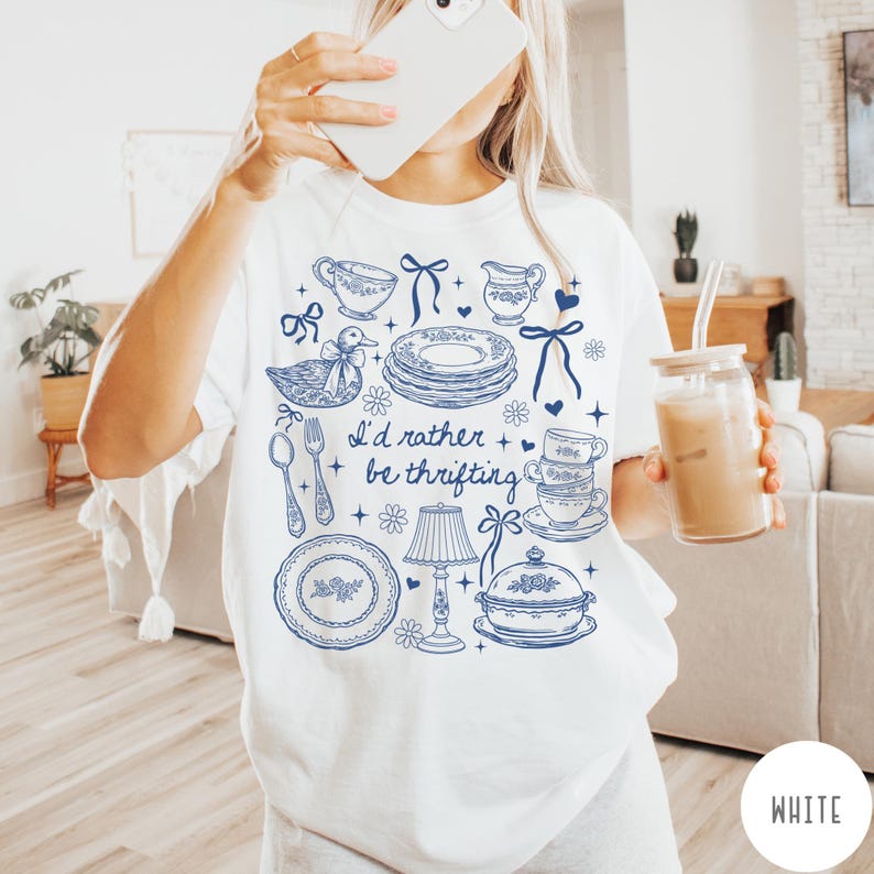 May include: White t-shirt featuring a blue illustration of vintage crockery, a duck, and the text "I'd rather be thrifting." The design includes plates, teacups, and a lamp. The shirt is a comfortable, casual style.