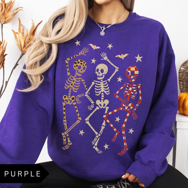 Puede incluir: Una sudadera morada con un gr&aacute;fico de tres esqueletos bailando en oro, rojo y blanco. El dise&ntilde;o incluye estrellas y murci&eacute;lagos, creando un tema festivo de Halloween. La palabra "PURPLE" est&aacute; en la parte inferior.