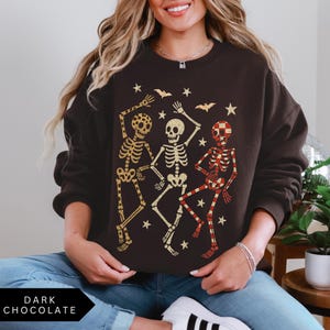 Puede incluir: Sudadera de color chocolate oscuro con un gr&aacute;fico de tres esqueletos bailando en dorado y rojo, con murci&eacute;lagos y estrellas. El texto "DARK CHOCOLATE" est&aacute; en la parte inferior. Un dise&ntilde;o casual y festivo para Halloween.