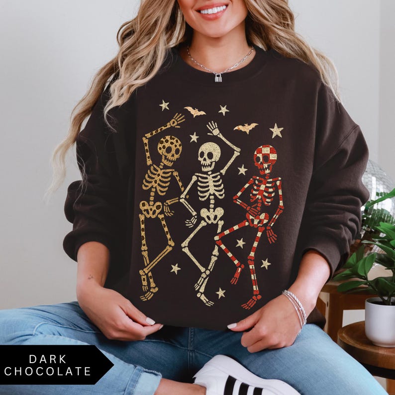 Puede incluir: Sudadera de color chocolate oscuro con un gr&aacute;fico de tres esqueletos bailando en dorado y rojo, con murci&eacute;lagos y estrellas. El texto "DARK CHOCOLATE" est&aacute; en la parte inferior. Un dise&ntilde;o casual y festivo para Halloween.