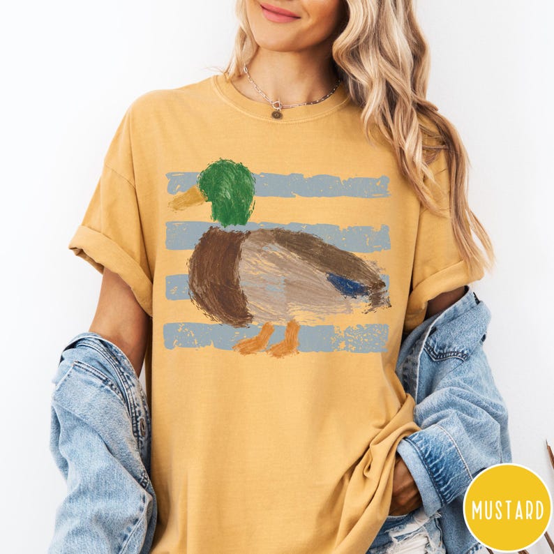 May include: A mustard yellow short-sleeve t-shirt featuring a graphic of a mallard duck with a green head and brown body, rendered in a textured, painterly style over horizontal light blue stripes. The shirt has a crew neckline and loose fit.