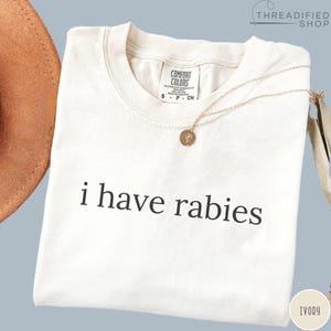 May include: Ivory-colored t-shirt with the phrase "i have rabies" printed in black. A gold necklace with a small pendant rests on the shirt. The shirt has a Comfort Colors tag at the neckline.