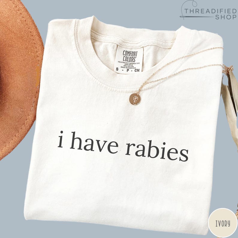May include: Ivory-colored t-shirt with the phrase "i have rabies" printed in black. A gold necklace with a small pendant rests on the shirt. The shirt has a Comfort Colors tag at the neckline.