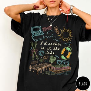 Puede incluir: Camiseta negra con un dise&ntilde;o gr&aacute;fico colorido con ilustraciones tem&aacute;ticas de lago. El dise&ntilde;o incluye una nevera port&aacute;til, gafas de sol, protector solar, un sol, nubes, un salvavidas, remos, una pelota de playa, peces, sandalias, un muelle y un barco. El texto dice "I'd rather be at the lake."