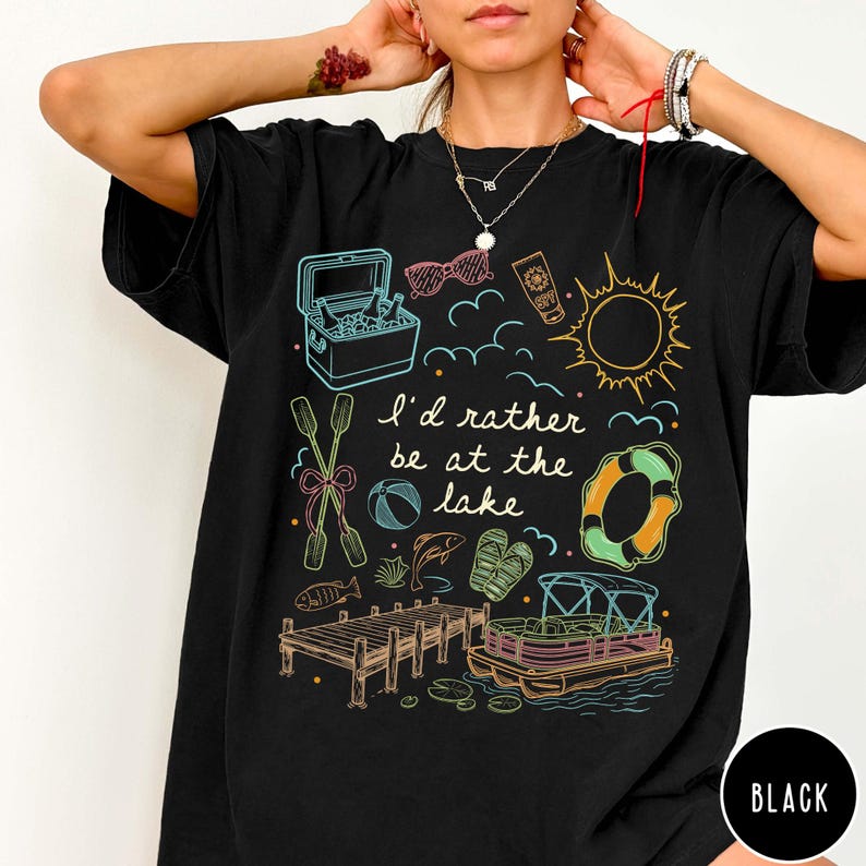 Puede incluir: Camiseta negra con un dise&ntilde;o gr&aacute;fico colorido con ilustraciones tem&aacute;ticas de lago. El dise&ntilde;o incluye una nevera port&aacute;til, gafas de sol, protector solar, un sol, nubes, un salvavidas, remos, una pelota de playa, peces, sandalias, un muelle y un barco. El texto dice "I'd rather be at the lake."