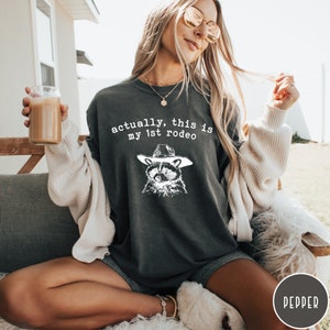 May include: A dark gray t-shirt with a white graphic of a raccoon wearing a cowboy hat. The text "actually, this is my 1st rodeo" is printed on the shirt.