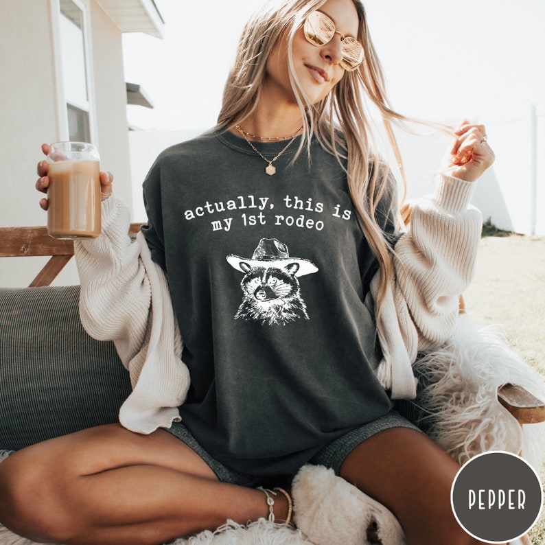 May include: A dark gray t-shirt with a white graphic of a raccoon wearing a cowboy hat. The text "actually, this is my 1st rodeo" is printed on the shirt.