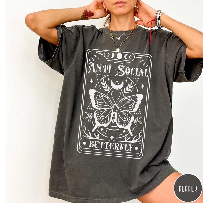 Puede incluir: Una camiseta de gran tama&ntilde;o de color gris oscuro con un gr&aacute;fico blanco de una carta de tarot con una mariposa y el texto "Anti-Social Butterfly".