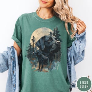May include: Light green t-shirt featuring a graphic of a black Labrador Retriever with a full moon and forest background. The design includes two images of the dog. The shirt has a crew neck and short sleeves. The words "LIGHT GREEN" are in a circle on the bottom right.