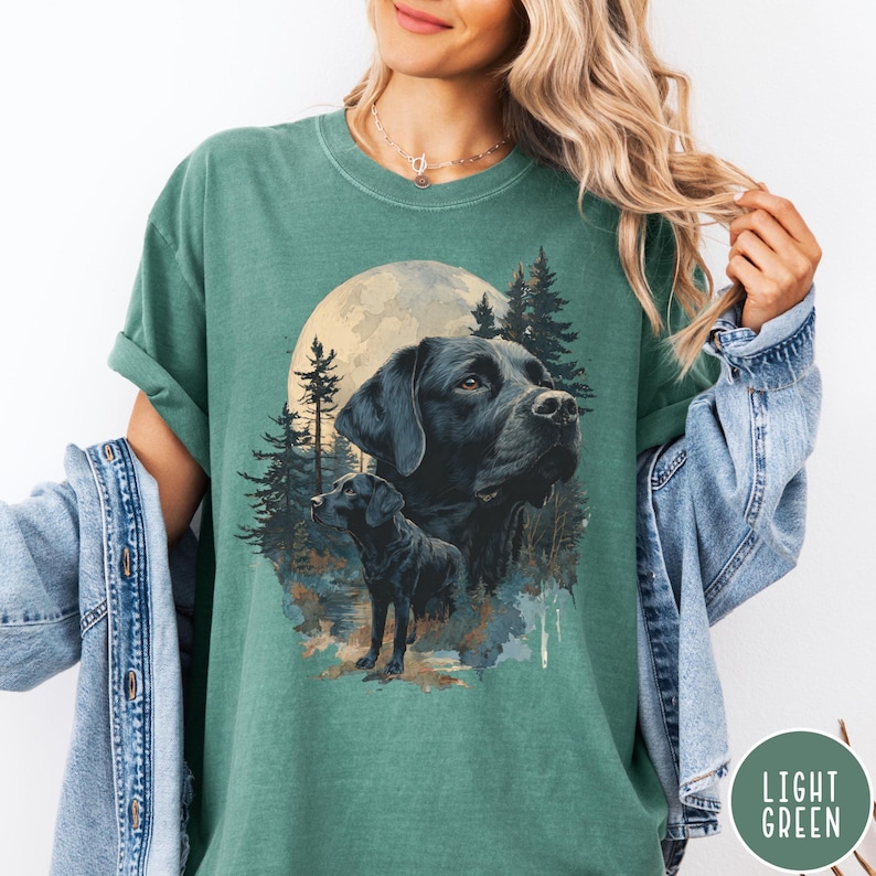 May include: Light green t-shirt featuring a graphic of a black Labrador Retriever with a full moon and forest background. The design includes two images of the dog. The shirt has a crew neck and short sleeves. The words "LIGHT GREEN" are in a circle on the bottom right.
