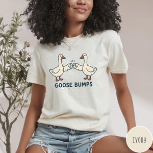 Puede incluir: Una camiseta de manga corta color marfil con un gr&aacute;fico de dos gansos blancos chocando los pu&ntilde;os. Debajo de los gansos, el texto GOOSE BUMPS est&aacute; impreso en letras may&uacute;sculas de color azul oscuro. La camiseta tiene cuello redondo.