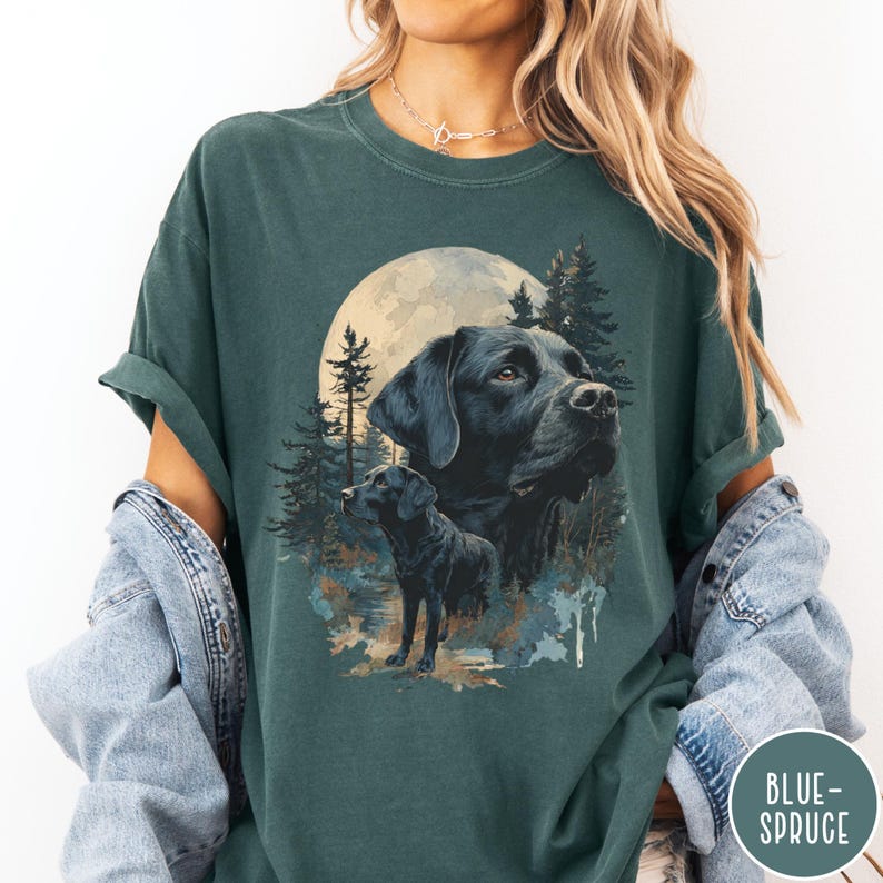 May include: Dark teal t-shirt featuring a graphic of a black Labrador Retriever with a full moon and forest background. The design includes two images of the dog, one large and one smaller, with a forest scene. The text "BLUE-SPRUCE" is in a circle on the bottom right.