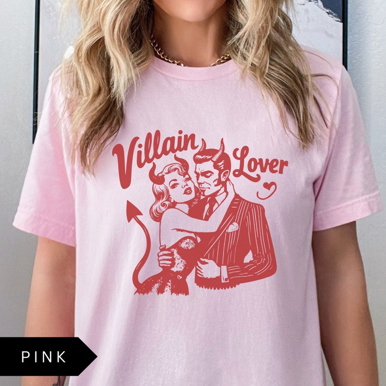 May include: Pink t-shirt with a graphic print of a woman with devil horns embracing a man in a suit. The text "Villain Lover" is printed above the graphic.