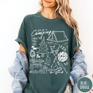 Może przedstawiać: Ciemnozielona koszulka z białymi grafikami o tematyce kempingowej. Projekt zawiera namiot, ognisko, drzewa oraz frazy &bdquo;Let's Go Camping&rdquo; i &bdquo;Adventure Awaits&rdquo;. Koszulka ma r&oacute;wnież słowa &bdquo;Happy Campers&rdquo; w okrągłym wzorze. W prawym dolnym rogu znajdują się słowa &bdquo;Blue Spruce&rdquo;.