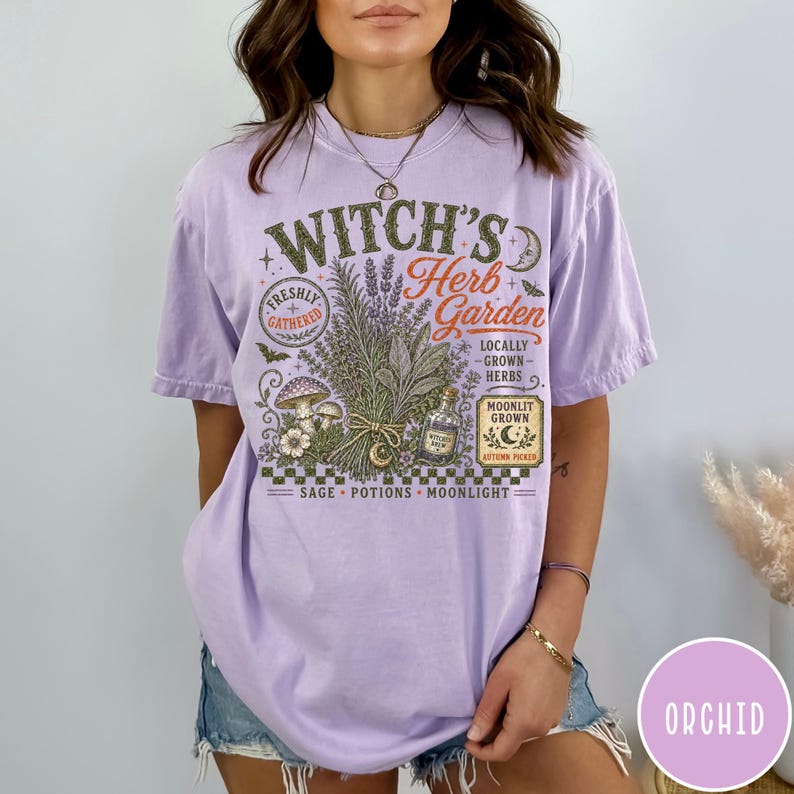 May include: A light purple short-sleeve t-shirt featuring a graphic print with the text Witch's Herb Garden, Freshly Gathered, Locally Grown Herbs, Moonlit Grown, Autumn Picked, Sage, Potions, Moonlight, and a bundle of herbs, mushrooms, and a bottle.