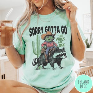 May include: Mint green t-shirt with the text "SORRY GOTTA GO THE VIBES ARE OFF." The graphic features a frog wearing a cowboy hat riding a raccoon, with a cactus in the background. The shirt is a casual, unisex style.