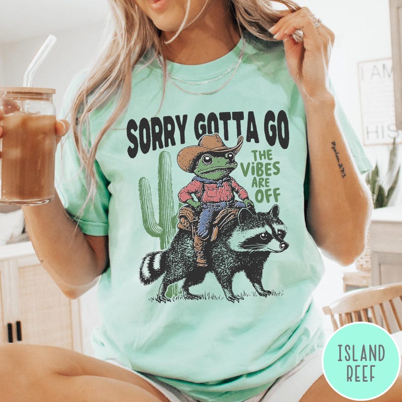 May include: Mint green t-shirt with the text "SORRY GOTTA GO THE VIBES ARE OFF." The graphic features a frog wearing a cowboy hat riding a raccoon, with a cactus in the background. The shirt is a casual, unisex style.