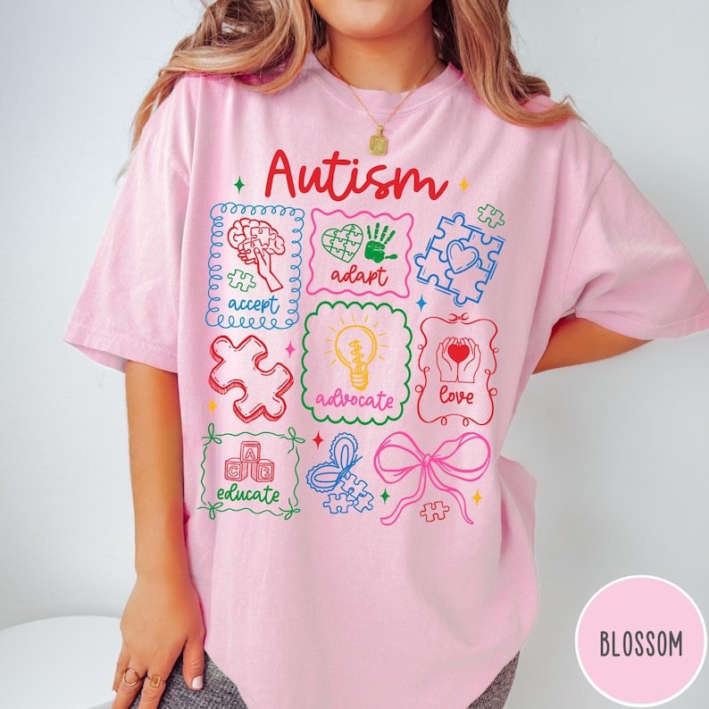 May include: Pink t-shirt with the word "Autism" in red script. The shirt features colorful illustrations of puzzle pieces, a brain, a heart, a handprint, a lightbulb, and a heart with the words "accept", "adapt", "advocate", "love", and "educate".