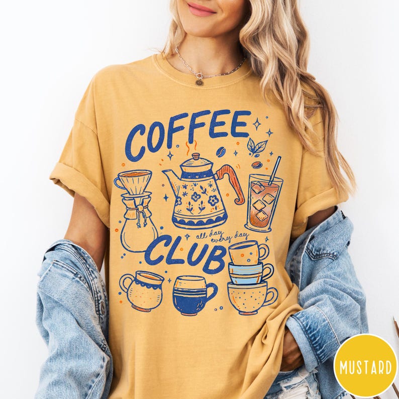 May include: Mustard yellow t-shirt with a blue and white "Coffee Club" graphic. The design includes coffee-related illustrations such as a teapot, coffee cups, and a glass of iced coffee. The shirt has short sleeves and a relaxed fit.