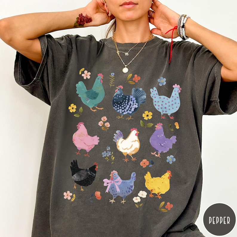 K&ouml;nnte beinhalten: Ein dunkelgraues T-Shirt mit einer bunten Illustration von H&uuml;hnern und Blumen. Die H&uuml;hner sind in verschiedenen Farben, darunter rosa, lila, blau, gr&uuml;n und wei&szlig;. Die Blumen sind in Rosa-, Blau- und Gelbt&ouml;nen gehalten.