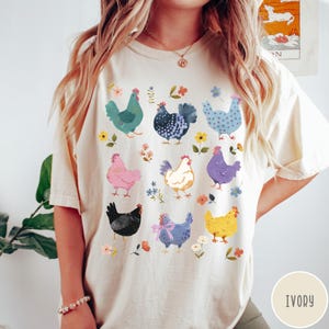 K&ouml;nnte beinhalten: Ein cremefarbenes T-Shirt mit einem bunten Grafikdruck von verschiedenen H&uuml;hnern. Die H&uuml;hner sind in verschiedenen Posen und Farben dargestellt, darunter rosa, blau, gelb und schwarz. Das Shirt hat einen Rundhalsausschnitt und kurze &Auml;rmel. Der Text "IVORY" ist in der rechten unteren Ecke des Bildes gedruckt.