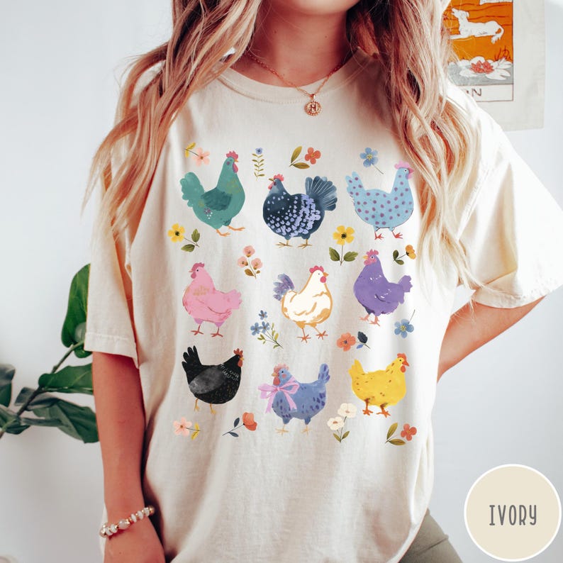 K&ouml;nnte beinhalten: Ein cremefarbenes T-Shirt mit einem bunten Grafikdruck von verschiedenen H&uuml;hnern. Die H&uuml;hner sind in verschiedenen Posen und Farben dargestellt, darunter rosa, blau, gelb und schwarz. Das Shirt hat einen Rundhalsausschnitt und kurze &Auml;rmel. Der Text "IVORY" ist in der rechten unteren Ecke des Bildes gedruckt.
