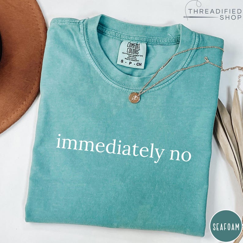 May include: Seafoam green t-shirt with the phrase "immediately no" in white block letters. The shirt has a crew neck and short sleeves. A gold necklace with a pendant rests on the shirt. The shirt is labeled "Comfort Colors".