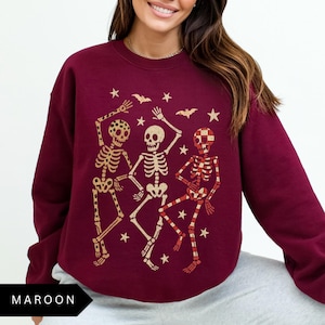 Puede incluir: Sudadera granate con una ilustraci&oacute;n dorada y roja de tres esqueletos bailando. El dise&ntilde;o incluye estrellas y murci&eacute;lagos, ideal para Halloween. La palabra "MAROON" est&aacute; impresa en la parte inferior. Prenda de temporada.