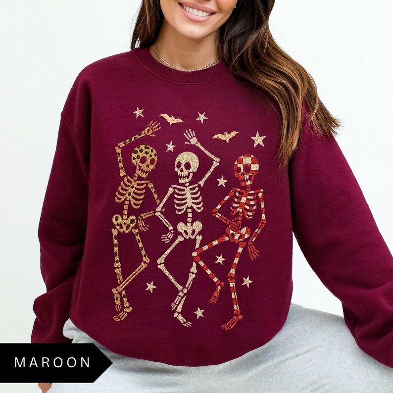 Puede incluir: Sudadera granate con una ilustraci&oacute;n dorada y roja de tres esqueletos bailando. El dise&ntilde;o incluye estrellas y murci&eacute;lagos, ideal para Halloween. La palabra "MAROON" est&aacute; impresa en la parte inferior. Prenda de temporada.