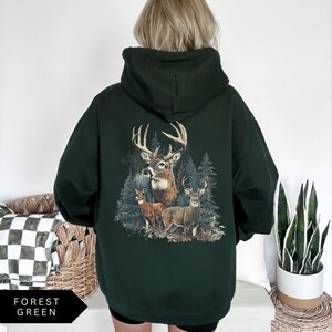 Op de afbeelding: Bosse groene hoodie met een hertenafbeelding op de rug. De afbeelding toont drie herten in een bosrijke omgeving. De hoodie heeft een capuchon en een relaxte pasvorm. De tekst "FOREST GREEN" staat linksonder.