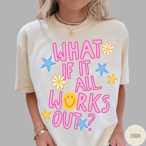 May include: An ivory short-sleeve t-shirt featuring the pink text WHAT IF IT ALL WORKS OUT? with a yellow smiley face replacing the letter O. The design includes scattered yellow daisies, blue stars, and yellow stars printed across the front.