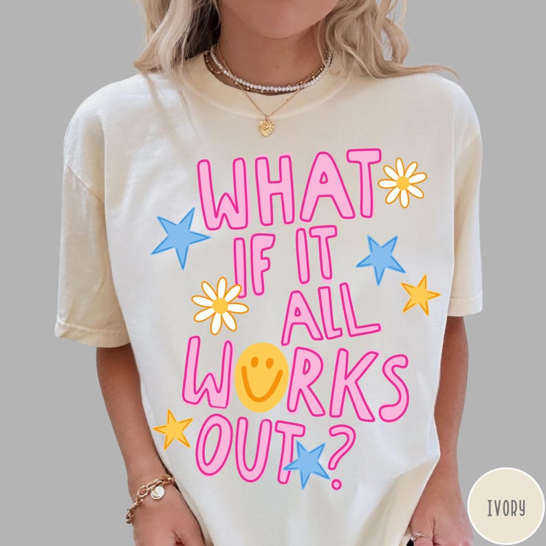 May include: An ivory short-sleeve t-shirt featuring the pink text WHAT IF IT ALL WORKS OUT? with a yellow smiley face replacing the letter O. The design includes scattered yellow daisies, blue stars, and yellow stars printed across the front.