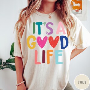 K&ouml;nnte beinhalten: Ein elfenbeinfarbenes Kurzarm-T-Shirt mit dem bunten Schriftzug "IT'S A GOOD LIFE" auf der Vorderseite. Die Buchstaben und zwei Herzformen sind in verschiedenen leuchtenden Farben wie Gr&uuml;n, Lila, Pink, Gelb, Rot, Blau und Orange dargestellt.