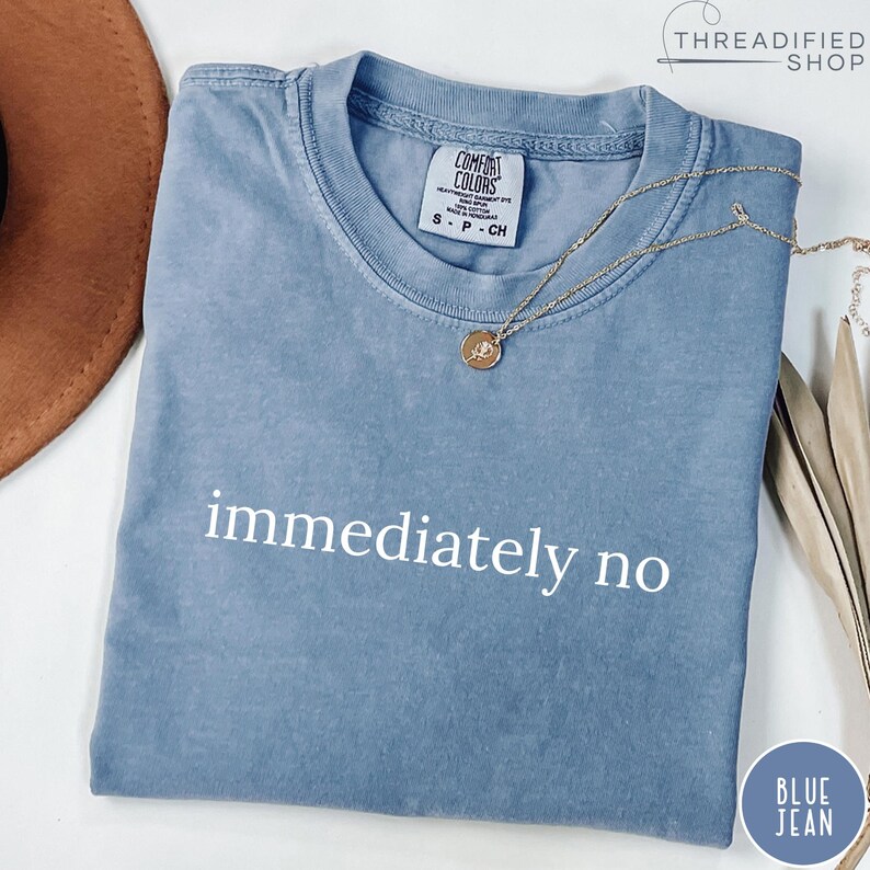 May include: A blue jean Comfort Colors t-shirt with the words "immediately no" printed in white. A gold necklace with a pendant rests on the shirt. The shirt is a light blue color.